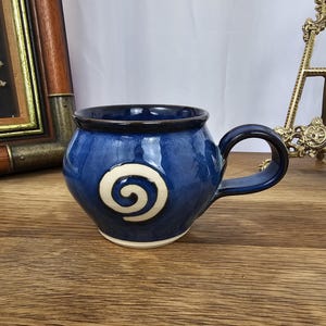 Può includere: Tazza in ceramica fatta a mano con smalto blu intenso e design a spirale bianco. La tazza ha una forma arrotondata, un bordo blu scuro e un manico robusto. Perfetta per gustare caffè o tè.