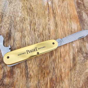 May include: A gold-colored pocket knife with a bottle opener and a blade. The knife is engraved with the words "MONTRES PIAGET WATCHES". The knife is on a wooden surface.