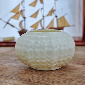 Può includere: Un piccolo vaso in ceramica bianco sporco con un motivo strutturato a forma di onda. Il vaso ha una forma rotonda e poggia su una superficie di legno. Sullo sfondo, un'immagine sfocata di un modello di veliero è visibile.