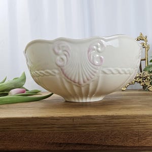 May include: A cream ceramic bowl with a scalloped edge and pink shell and scroll patterns. The bowl is on a wooden surface, with green leaves and a pink flower to the left. The bowl is likely for decorative use.