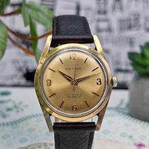 Könnte beinhalten: Eine goldfarbene Vintage-Armbanduhr mit einem schwarzen Lederarmband. Das Zifferblatt zeigt den Markennamen "BETINA" und den Text "17 RUBIS INCABLOC". Die Uhr hat goldene Zeiger und Stundenmarkierungen, mit den Zahlen 3, 6, 9 und 12.