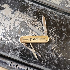 May include: A gold-toned multi-tool pocket knife featuring the text MONTRES PIAGET WATCHES on the handle. The tool includes two extended metal blades and a pair of small metal scissors, all displayed in an open position with a metallic finish.
