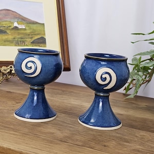 Può includere: Due calici in ceramica blu con un disegno a spirale bianco. I calici hanno una base svasata e un bordo blu scuro. Sono esposti su una superficie di legno. I calici sono alti circa 10 cm.