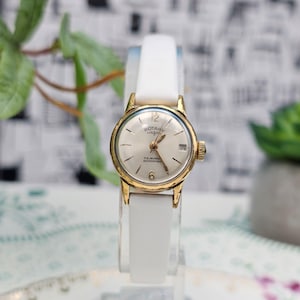 May include: A vintage gold-plated Rotary watch with a white strap. The watch face is silver with gold hands and hour markers. The watch has a date window at the 3 o'clock position. The watch is displayed on a clear stand.
