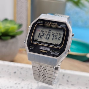 May include: A silver-toned Darwil digital watch displaying the time 12:07:52. The watch has a black face with white text, including the day and date. The watchband is a matching silver metal link style.