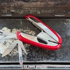 Op de afbeelding: Een rood Wenger Swiss Business Tool multifunctioneel gereedschap met een zilverkleurige metalen afwerking. Het apparaat heeft verschillende uitgeklapte roestvrijstalen onderdelen, waaronder een schaar, een mes en een vijl. De tekst SWISS BUSINESS Tool by WENGER staat op de zijkant gedrukt.