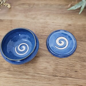 Può includere: Un set fatto a mano composto da una ciotola e un coperchio in ceramica blu. La ciotola rotonda e il coperchio circolare presentano un motivo a spirale bianco. Il set è posto su una superficie di legno.