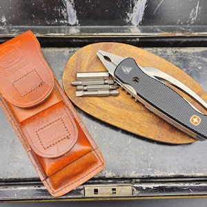 Op de afbeelding: Een zwart multi-tool met een getextureerde handgreep en zilveren mes, samen met een set zilveren bits, ligt op een houten oppervlak. Een bruine leren etui met het "Wenger"-logo is ook aanwezig. De tool heeft een rood-wit kruis embleem.