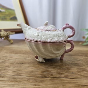 Puede incluir: Pequeña caja de baratijas decorativa con forma de tetera. La pieza de cerámica es blanca con una superficie texturizada y detalles en rosa. La tapa y el asa son rosas, y la base tiene tres patitas. La tetera está sobre una superficie de madera.