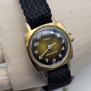 Könnte beinhalten: Vintage Timex-Uhr mit goldfarbenem Gehäuse und schwarzem geflochtenem Armband. Das Zifferblatt ist dunkel mit weißen Ziffern und Zeigern. Auf dem Zifferblatt steht "Timex" und "Water Resistant".