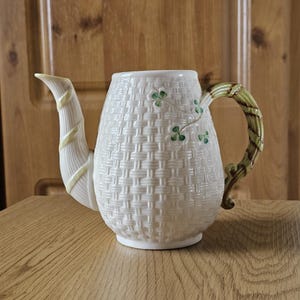 Puede incluir: Una tetera de cerámica color crema con un patrón de tejido de cesta texturizado en el cuerpo. El pico está envuelto en un detalle de cinta amarilla pálida, mientras que el asa tiene forma de enredadera verde. Pequeños motivos de tréboles verdes decoran el lateral.
