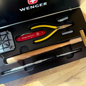 Op de afbeelding: Een Wenger horlogereparatieset in een zwarte presentatiedoos. De set bevat een rood zakmes met de tekst Wenger Watch S.A., een tang met gele handvatten, een koperen hamer met houten handvat, een grijs blok en twee metalen precisiegereedschappen.