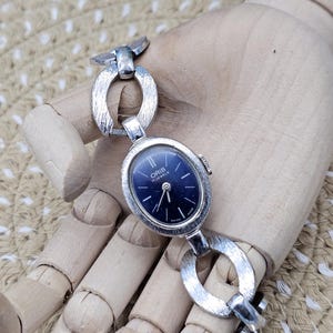 Puede incluir: Reloj de pulsera Oris vintage plateado con esfera ovalada azul oscuro. El reloj presenta un diseño de pulsera de eslabones. La esfera del reloj muestra el nombre de la marca y "17 Jewels". El reloj se muestra en una mano de madera.
