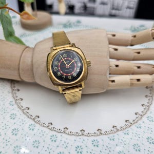 May include: A vintage gold-toned watch with a black face and red accents. The watch has a rectangular case and a gold mesh band. The watch is displayed on a wooden hand. The watch face displays the brand name "KEN".