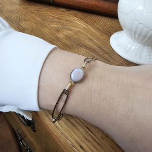 May include: A delicate gold-toned bracelet featuring an oval, opalescent gemstone. The bracelet has a unique design with elongated, rectangular links and is worn on a wrist with a white sleeve.