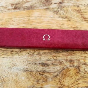 Pode incluir: Uma caixa retangular, cor de vinho, com um símbolo Omega branco na parte superior. A caixa é feita de um material texturizado e está colocada sobre um fundo de madeira. A caixa é provavelmente para um relógio ou joia.