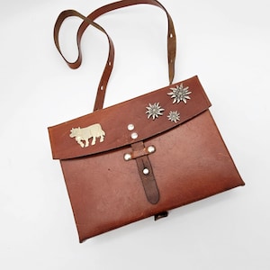 May include: Brown leather shoulder bag with a flap closure and adjustable strap. The bag features a metal cow emblem and three star-shaped embellishments on the flap. The bag has a buckle closure and is likely vintage.