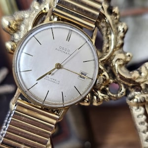 May include: A vintage OSCO automatic wristwatch with a gold-toned case and band. The watch face is silver with black hour markers and the text "OSCO AUTOMATIC 25 JEWELS SHOCKPROOF". The watch is displayed on a gold-colored decorative stand.