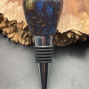 May include: A decorative bottle stopper with a dark blue and brown resin top, featuring gold accents. The stopper has a stainless steel base and black rubber rings. The product is designed for wine bottles.