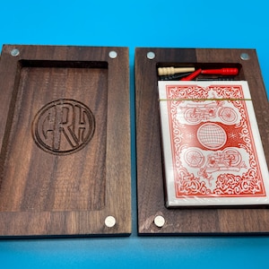 May include: Open wooden box with a deck of cards and small accessories. The box is dark brown with a recessed area and a carved circular design. The deck of cards has a red and white design with images of motorcycles. The box is open on a blue surface.