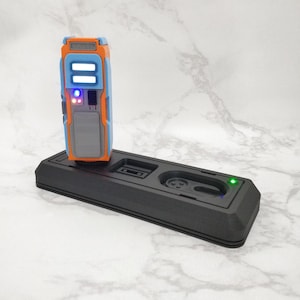 Op de afbeelding: Een zwart laadstation met een blauw, oranje en grijs apparaat dat is aangesloten. Het apparaat heeft twee witte lampjes en een rood lampje aan de voorkant. Het laadstation heeft een groen lampje aan de voorkant.
