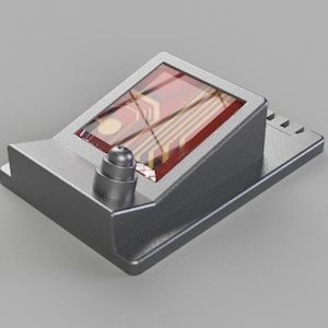 Puede incluir: Un pequeño dispositivo electrónico rectangular gris con una ventana transparente roja y dorada en la parte superior. La ventana muestra una placa de circuito con trazos rojos y dorados.