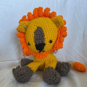 Op de afbeelding: Een gehaakt leeuwen speelgoed met een geel lichaam, een oranje manen en grijze poten. De leeuw heeft zwarte ogen en een zwarte neus.