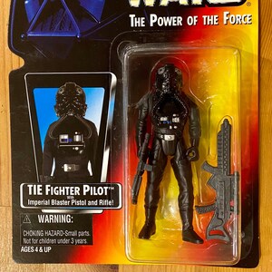 May include: A Star Wars action figure of a TIE Fighter Pilot in black and gray with a black helmet and a blaster rifle. The packaging is red, yellow, and black with the words "Star Wars" and "The Power of the Force".
