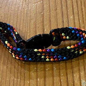 May include: A black woven bracelet with a colorful pattern of orange, yellow, blue, and purple threads. The bracelet has a black plastic clasp. The bracelet is approximately 0.75 inches wide.