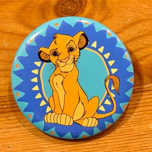 May include: A round button featuring a cartoon lion cub in shades of orange and yellow, set against a blue background. The button has a turquoise center and a decorative blue border with yellow accents. The lion cub is smiling.