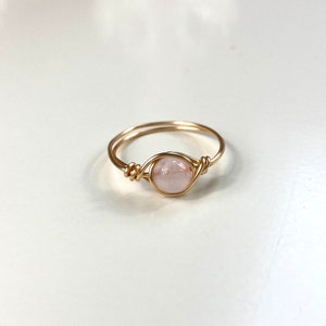 Könnte beinhalten: Ein zarter Ring mit einem hellrosa, runden Edelstein, der in einem goldfarbenen Drahtrahmen gefasst ist. Der Draht ist kunstvoll um den Stein gewickelt. Der Ring ist vor einem weißen Hintergrund platziert.