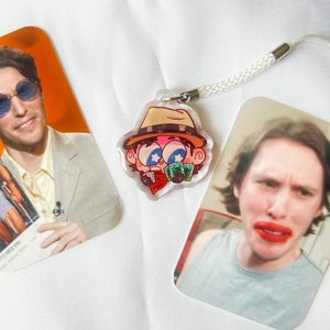 May include: A cartoon character with a brown hat, red scarf, and blue eyes is holding a stack of playing cards. The character is printed on a clear acrylic charm with a white lanyard.
