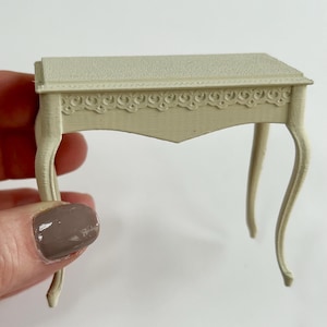 Pode incluir: Uma pequena mesa branca adornada com um design entalhado na parte superior e pernas curvas. A mesa é feita de um material liso e texturizado.