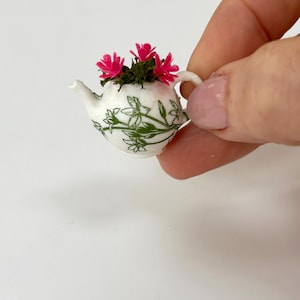 Puede incluir: Una tetera miniatura blanca con un estampado botánico verde de hojas y tallos, rellena de pequeñas flores de color rosa vibrante. La tetera tiene un asa curva y un pico corto, diseñada como pieza decorativa para casas de muñecas.