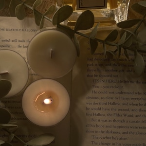 Op de afbeelding: Drie witte theelichtjes met een warme gloed. De kaarsen staan op een boek met tekst over de Doodsmerte uit de Harry Potter-serie.