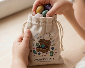 Könnte beinhalten: Ein beigefarbener Stoffbeutel mit Kordelzug und einem aufgedruckten Motiv einer offenen Schatztruhe, gefüllt mit bunten Edelsteinen, Sternen und Herzen. Unter der Illustration stehen der Text ZAUBERSCHATZ sowie eine Internetadresse.