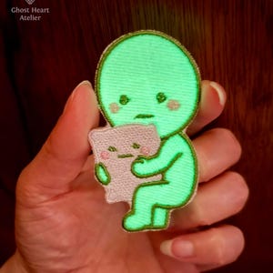 May include: An embroidered patch featuring a glowing green character with a round head and small facial features, holding a light pink rectangular pillow. The character has a sad expression and pink cheeks. The text Ghost Heart Atelier is visible.