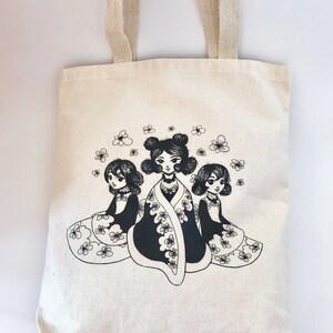 May include: A white canvas tote bag with black and white illustrations of three women with flowers. The women are wearing dresses and have flowers in their hair.