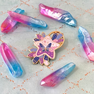 May include: A gold-tone enamel pin featuring a purple and pink star with a butterfly inside. The star is surrounded by crystals and a crescent moon. The pin is surrounded by iridescent pink and blue crystals.