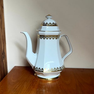 Könnte beinhalten: Weiße Porzellan-Kaffeekanne mit goldenen Verzierungen und geometrischem Muster. Die Kanne hat einen Deckel mit einem Knopf oben.