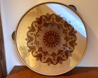 Op de afbeelding: Een ronde spiegel met een gouden lijst en een bruin kanten ontwerp in het midden. De spiegel staat op een houten oppervlak.