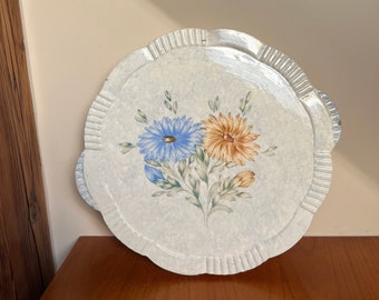 Op de afbeelding: Een wit keramiek bord met een gegolfde rand heeft een bloemenmotief met twee bloemen. Eén bloem is blauw en de andere is oranje.
