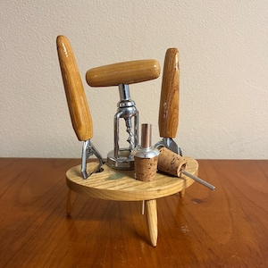 Può includere: Un set di apribottiglie in legno con un cavatappi, un apribottiglie e un tappo per bottiglie. Il set è su un piccolo supporto in legno con tre gambe.