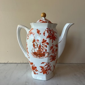 May include: White porcelain teapot with a lid and a handle. The teapot is decorated with a red floral and architectural pattern. The lid has a wooden knob and the spout and handle have gold trim.