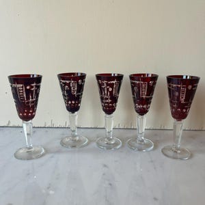 Pode incluir: Cinco taças de vinho de cristal vermelho rubi com hastes e bases transparentes. Os copos têm um design gravado detalhado. Os copos estão dispostos em fila sobre uma superfície de mármore branco.