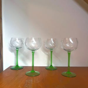 Könnte beinhalten: Vier klare Weingläser aus Glas mit grünen Stielen. Die Gläser sind in einer Reihe auf einer Holzoberfläche angeordnet.