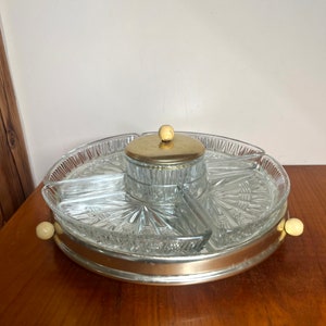 Może przedstawiać: Vintage szklana taca do serwowania z obrotową podstawą i pokrywką. Taca ma cztery sekcje i centralny przedział z pokrywką. Taca wykonana jest z przezroczystego szkła z srebrną obwódką i ma złotą pokrywkę z białym guzikiem.