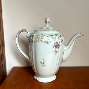 May include: A white porcelain teapot with a floral pattern and gold accents. The teapot has a lid with a knob and a spout. The teapot is sitting on a wooden surface.