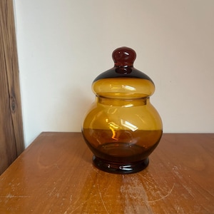 Könnte beinhalten: Eine bernsteinfarbene Glasdose mit einem dunkelroten Deckel. Die Dose hat einen runden Körper und eine breite Öffnung.