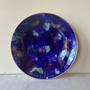 Op de afbeelding: Een ronde keramische plaat met een diepblauw en goud ontwerp. De plaat heeft een gouden rand en een gestructureerd, abstract patroon in tinten blauw, groen en goud. De plaat is een decoratief item.